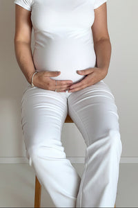 A pregnant woman sitting on a wooden stool, wearing a white ribbed camisole tank top and matching white pants against a plain off-white wall.