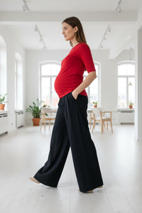 Woman in a red top and black pants standing in a bright, white room with large windows.